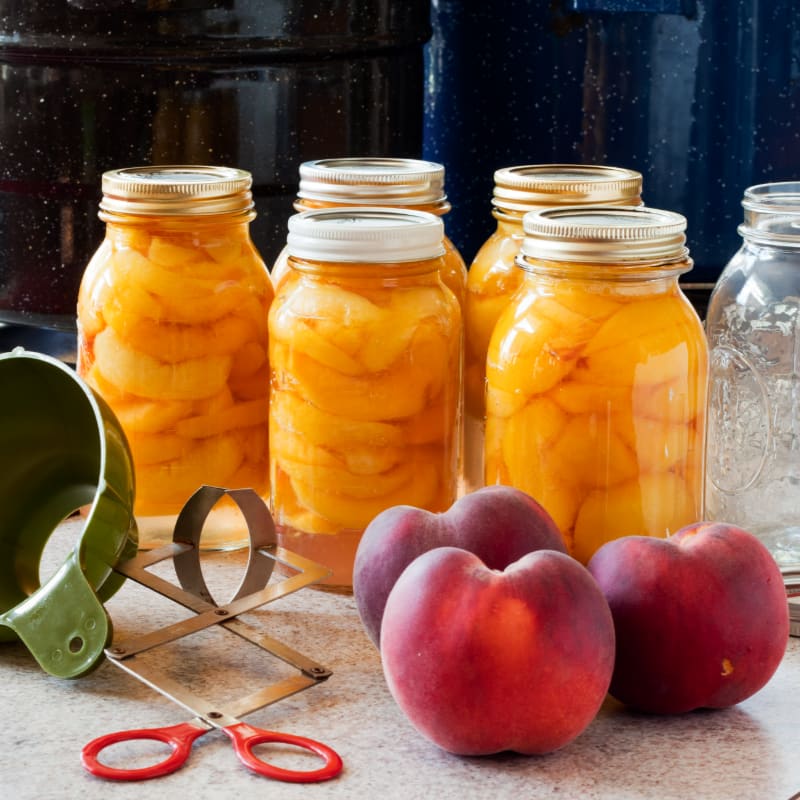 In the foreground, fresh peaches and canning tools sit on a counter, while in the background, jars of preserved peaches are lined up.