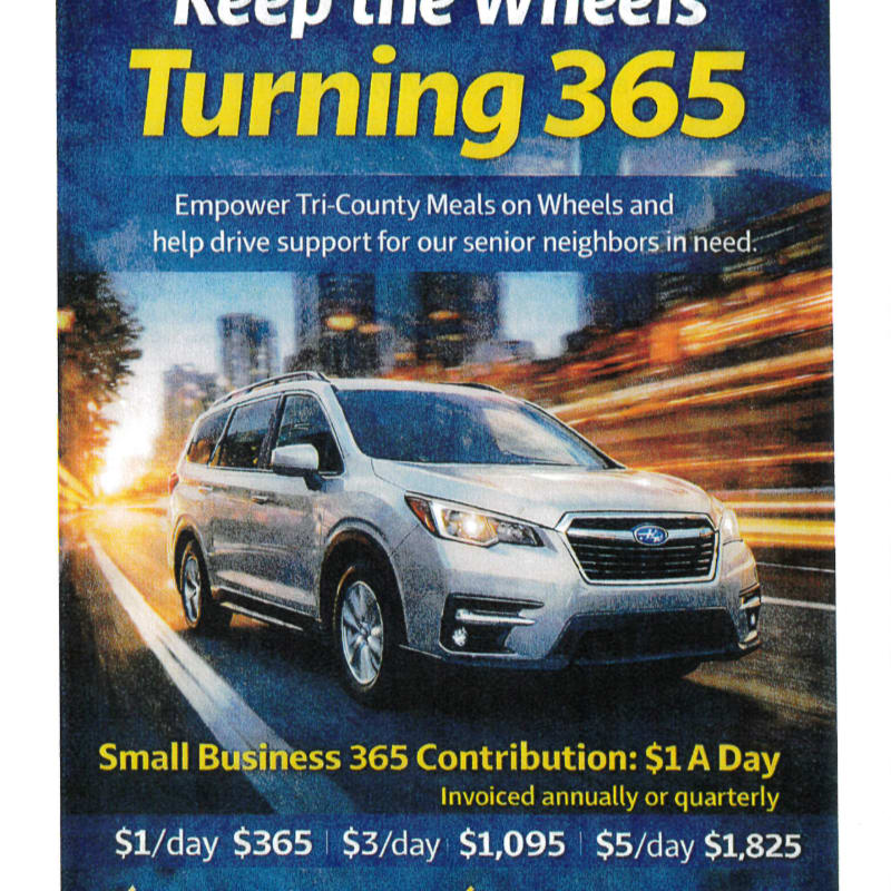 A silver Subaru SUV drives down a blurred road with a city skyline in the background, under a blue and yellow banner promoting "Keep the Wheels Turning 365" for Tri-County Meals on Wheels.