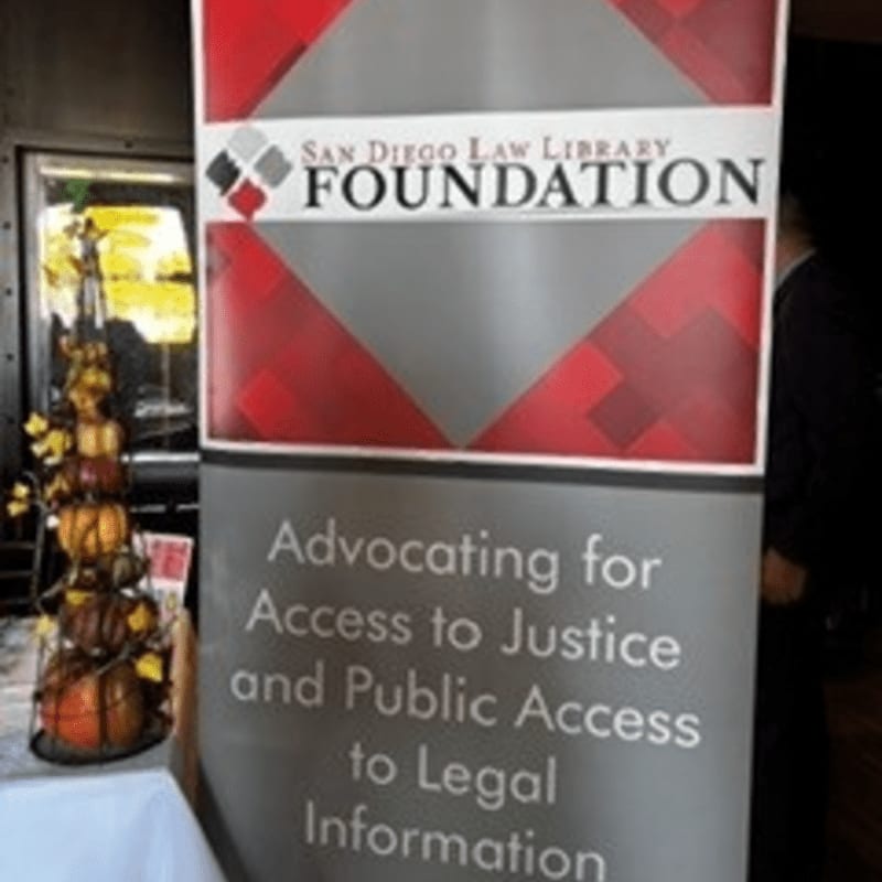 A banner for the San Diego Law Library Foundation stands in the foreground, with a decorative tiered display to its left and a blurred figure in a suit in the background.