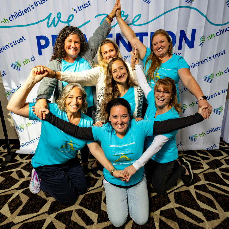 Six smiling women in teal shirts form a human pyramid in front of a banner that reads "we are PREVENTION."