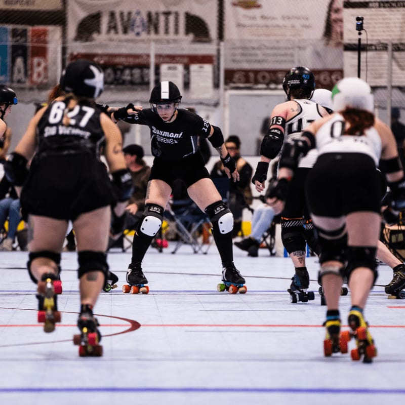 Four roller derby skaters in purple and teal uniforms jostle for position on a wooden floor, with a blurred background of a gymnasium.