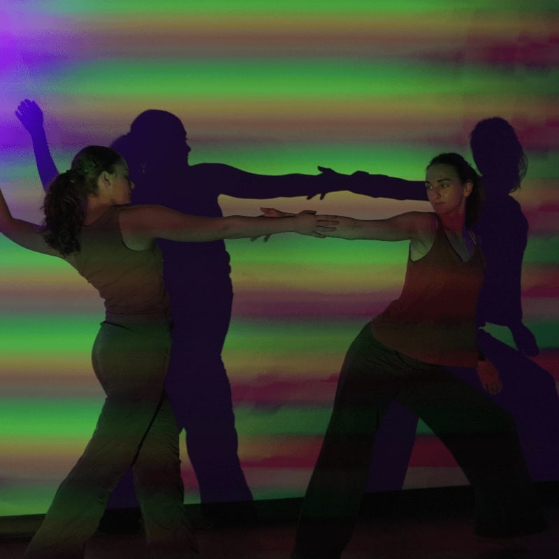 Two women in the foreground reach for each other's hands with colorful, streaky lights and shadows forming the background.