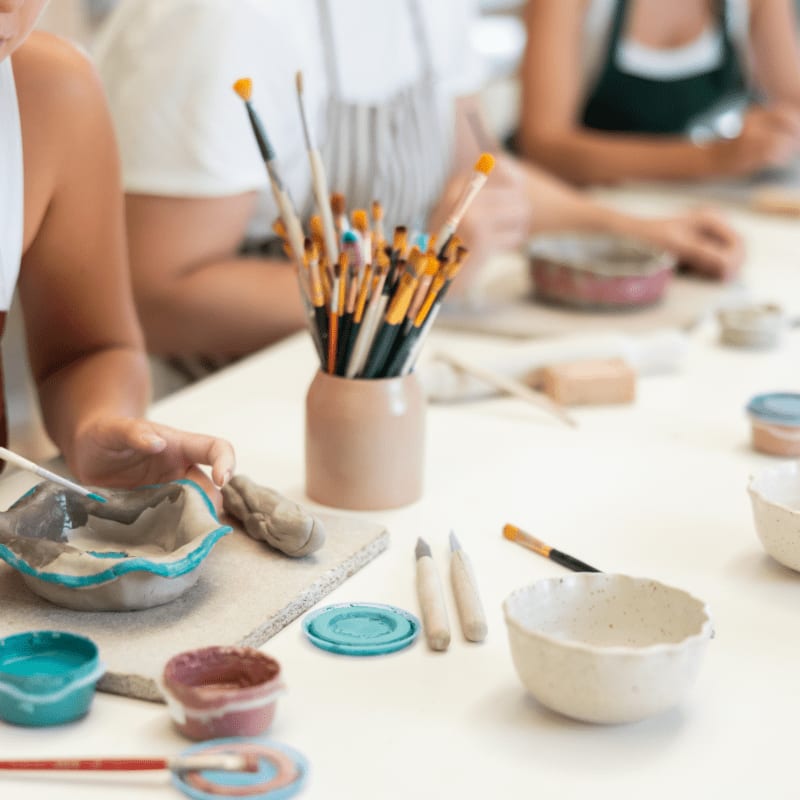 In the foreground, hands are working with clay and paints, while in the background, people are engaged in a pottery class.