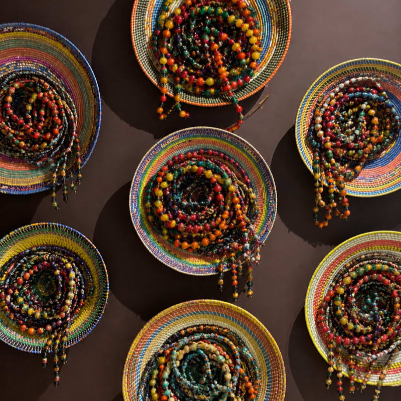 Colorful beaded necklaces are coiled in woven, circular baskets against a dark brown background.