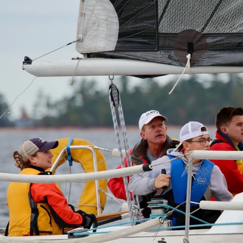 Four people in life vests are sailing on a boat with a blurry background of trees and water.