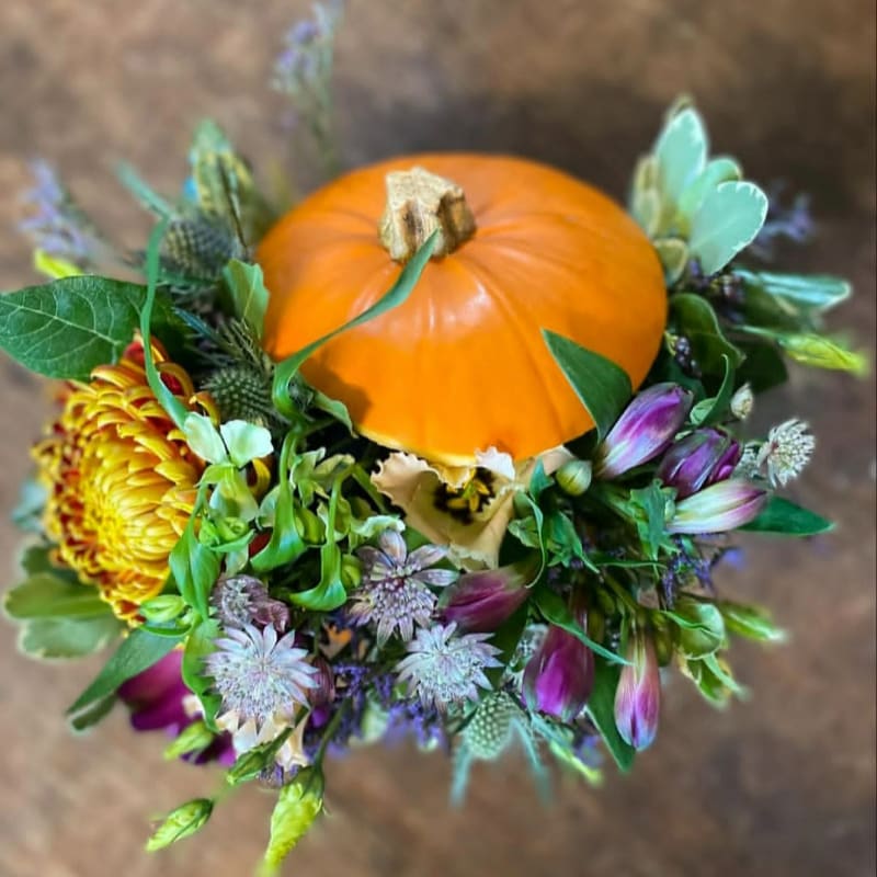 A small orange pumpkin sits atop a vibrant arrangement of fall flowers and greenery, set against a blurred wooden background.