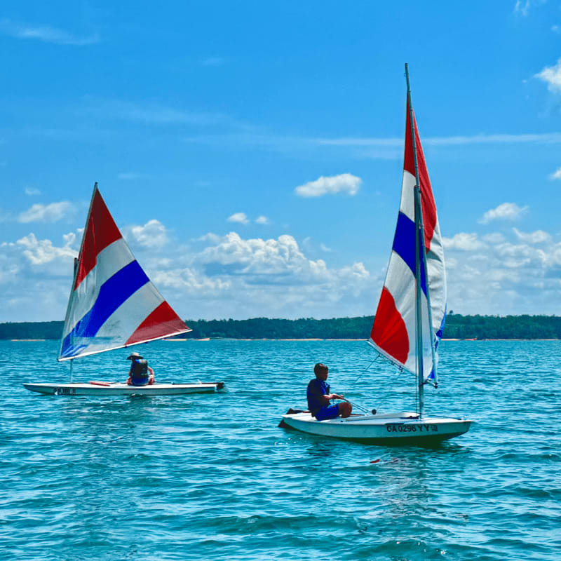 Two sailboats with red, white, and blue sails glide across a bright blue lake under a partly cloudy sky, with a tree-lined shore visible in the distance.