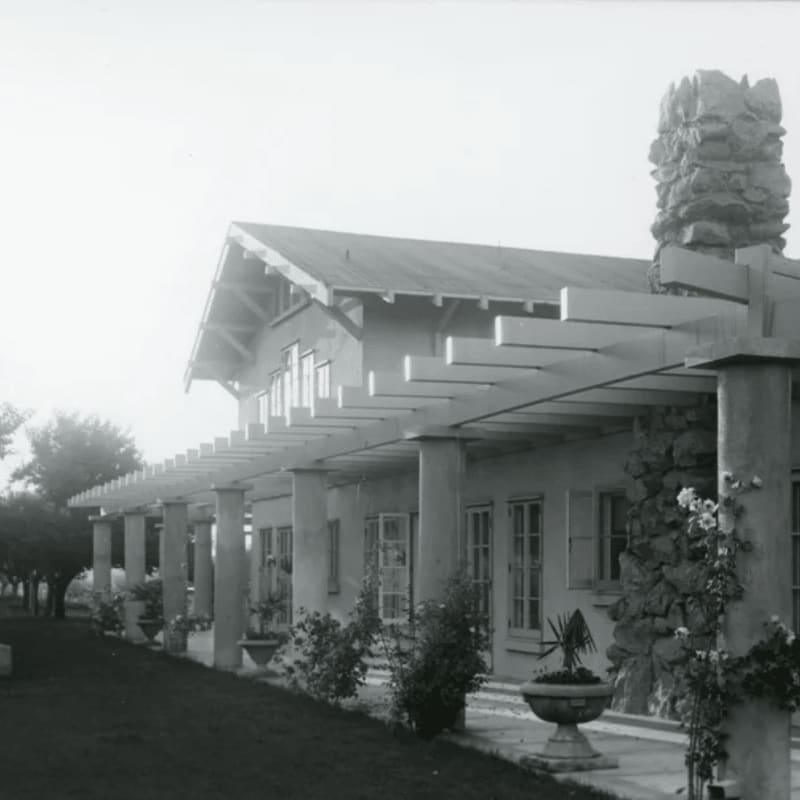 A long, covered porch with a stone chimney extends from a house with a sloped roof, while a grassy lawn and trees are visible to the left.