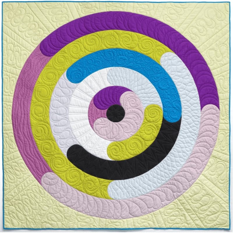A vibrant, concentric circular quilt design in purple, yellow, blue, white, pink, and black is set against a pale yellow background with intricate quilting patterns.