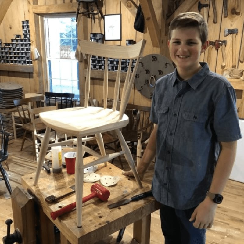 A smiling boy stands next to a newly crafted wooden chair on a workbench, with woodworking tools and supplies visible in the rustic workshop background.