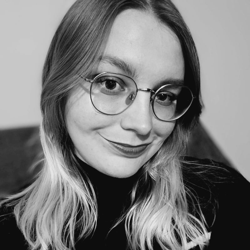 A young woman with blonde hair and glasses smiles at the camera in the foreground, with a blurred, dark background behind her.