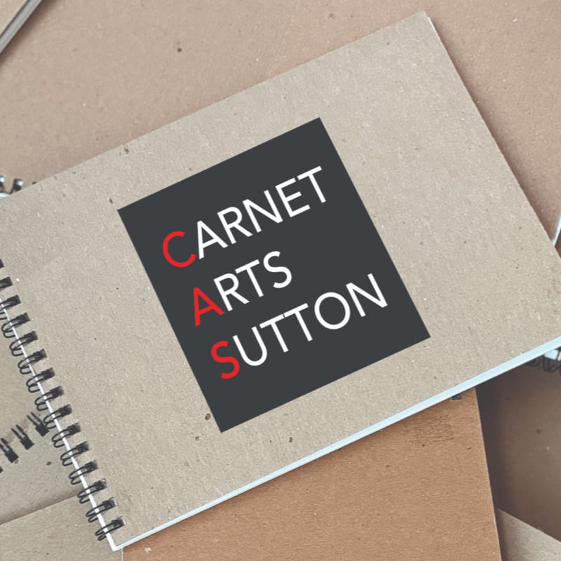 A spiral-bound notebook with a dark square logo containing white text "CARNET ARTS SUTTON" rests on a textured brown surface.