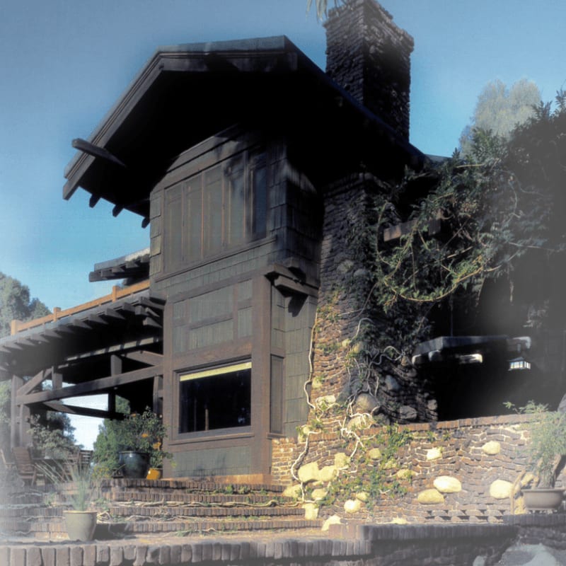 A dark, multi-story craftsman-style house with a prominent stone chimney and a wide porch is set against a backdrop of lush greenery and a clear blue sky.