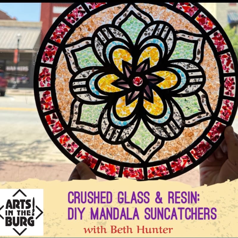A colorful, circular mandala suncatcher made of crushed glass and resin is held up against a blurred background of a street scene.