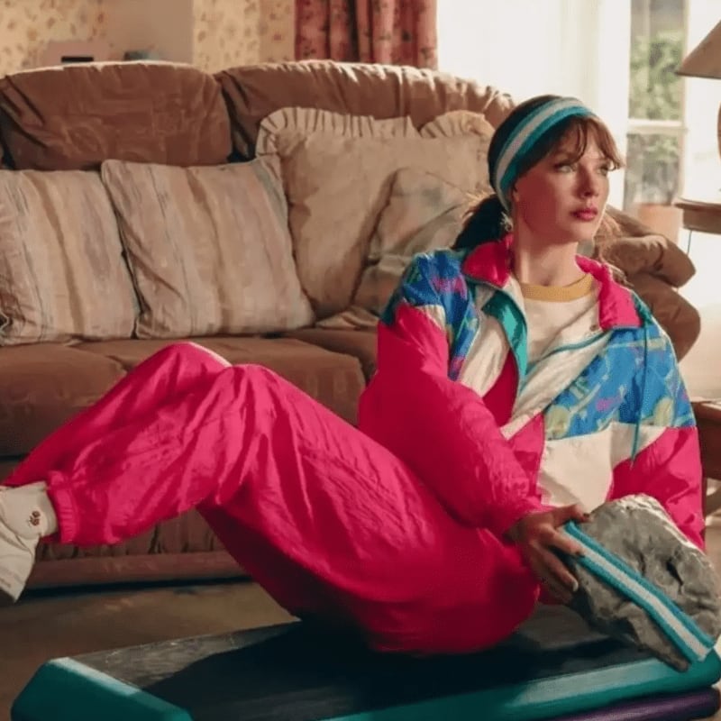 A woman in a vibrant tracksuit exercises on a step in a retro living room with a brown couch.