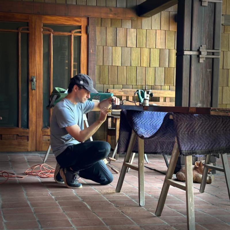 A man in a cap works on a wooden table supported by sawhorses covered in blue padding, with a wooden-shingled wall and glass doors in the background.