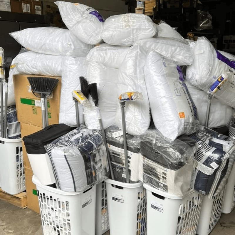 In the foreground, white laundry baskets are filled with bagged bedding and cleaning supplies, while the background is a cluttered storage area with stacked boxes and more bagged items.