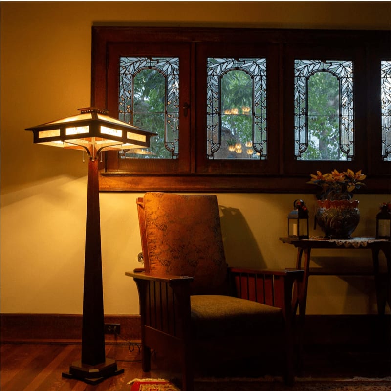 A tall, illuminated lamp stands in the foreground next to a wooden armchair, with a stained-glass window and a small table with flowers in the background.