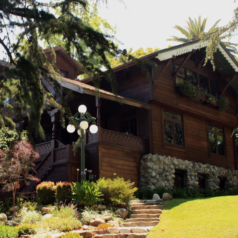A grand wooden house with a stone foundation is surrounded by lush greenery and trees, with a stone pathway leading up to its entrance.