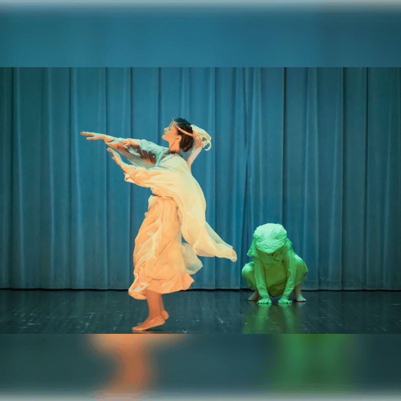 A dancer in a flowing costume poses on a stage with a green frog costume to her right, against a backdrop of blue curtains.