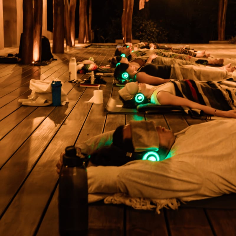 People lie on yoga mats on a wooden deck at night, wearing headphones with glowing green lights, while a dimly lit structure with wooden pillars stands in the background.