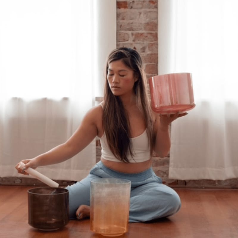 A woman in the foreground sits cross-legged with crystal singing bowls, while a brick wall and sheer curtains form the background.