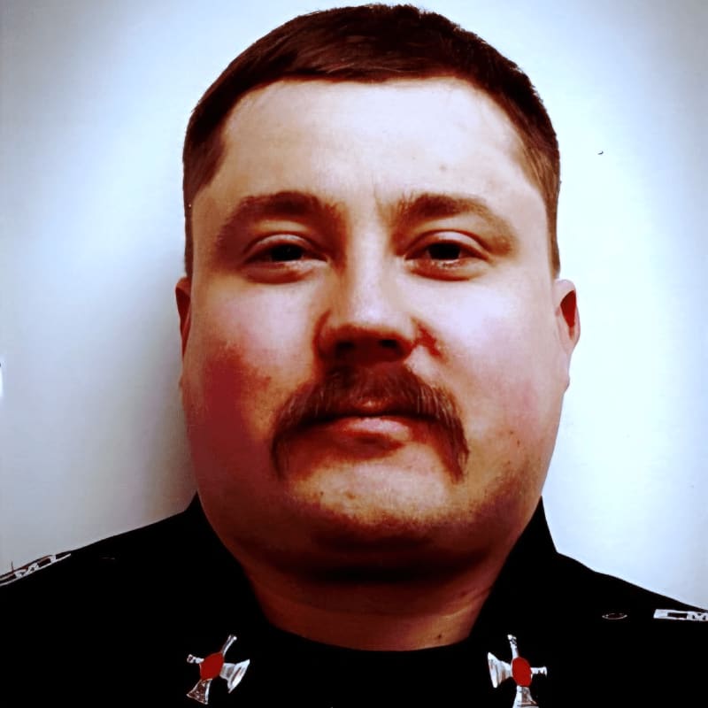 A man with a prominent mustache and a dark uniform with red and silver insignia is centered against a softly lit, plain background.