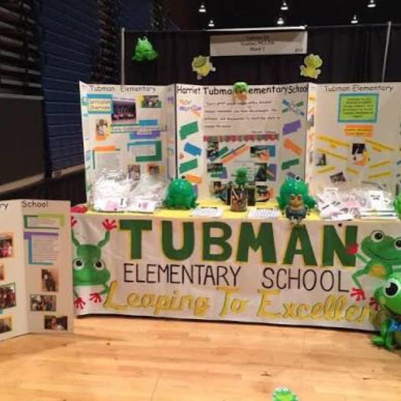 In the foreground, a table displays a "Tubman Elementary School" banner with frog decorations, while the background features display boards and black curtains.
