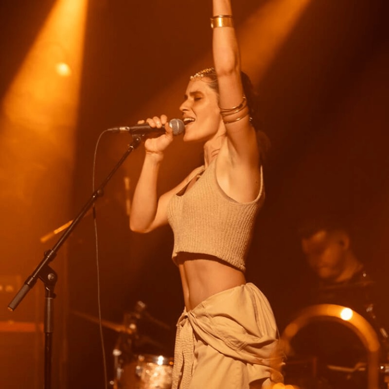 A woman in a crop top and flowing pants sings into a microphone on a stage, with warm stage lights illuminating the scene and blurred figures of band members in the background.
