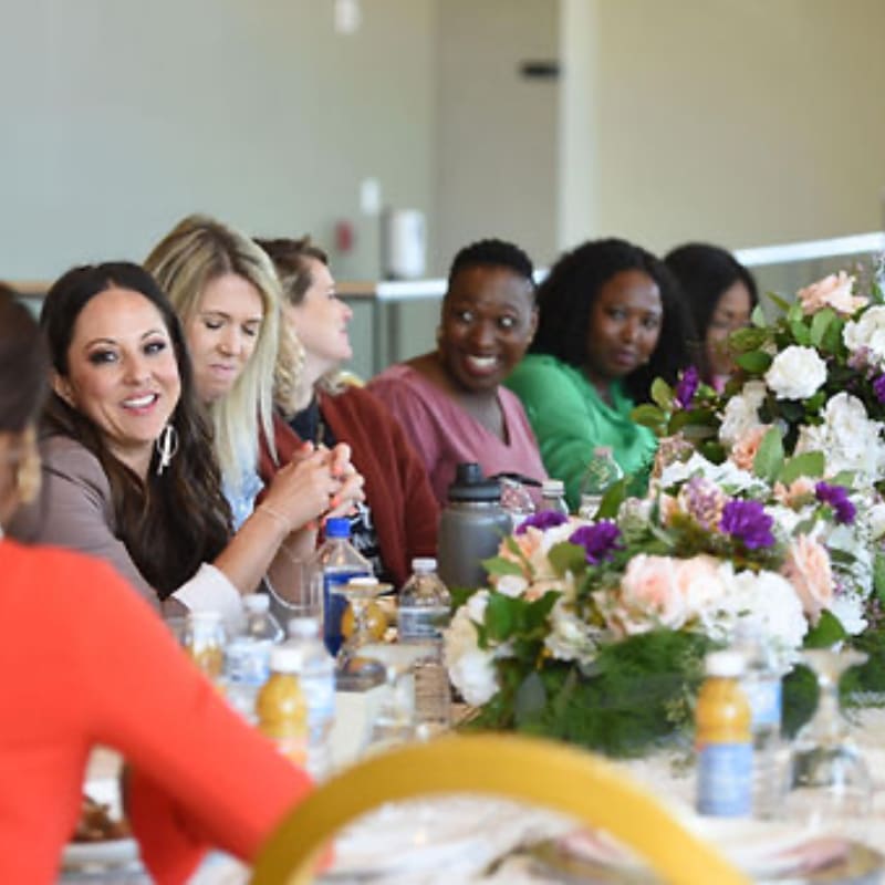 A group of women smile and chat around a table adorned with floral arrangements, with a bright, airy room in the background.