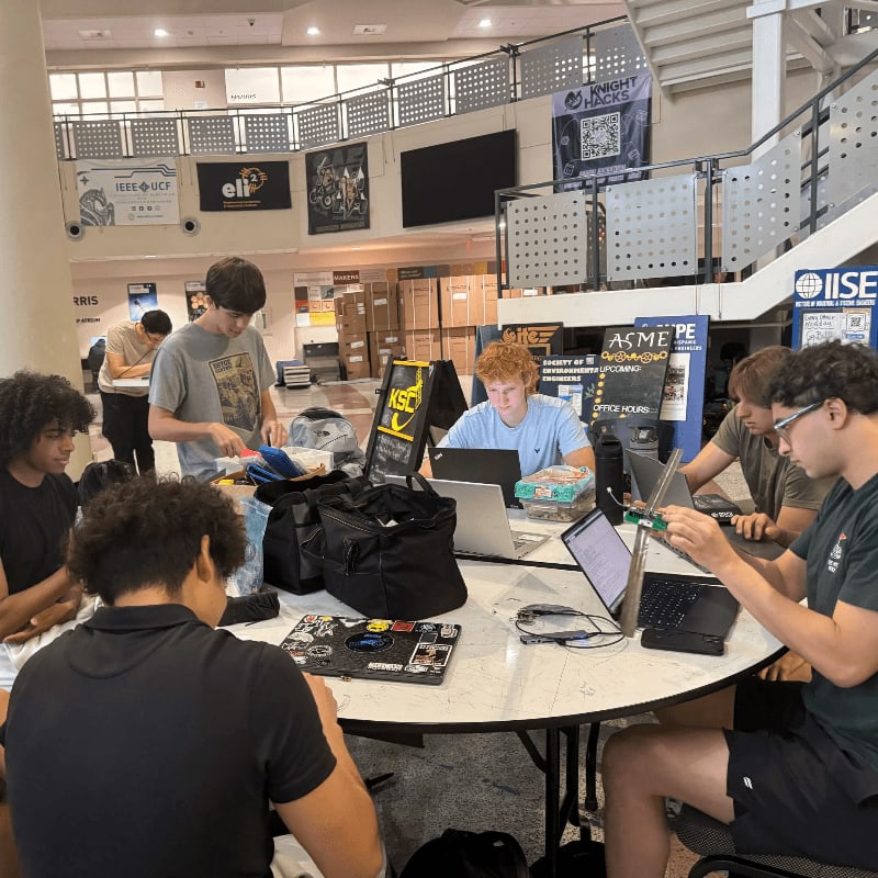 Several young men work on laptops around a large table in the foreground, while a multi-level atrium with banners and windows forms the background.