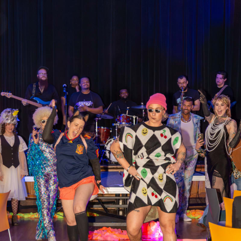 A group of performers in colorful costumes dance on a stage in front of a band playing instruments.