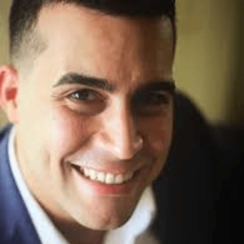 A man with dark hair and a bright smile is in the foreground, with a softly blurred, warm-toned background.