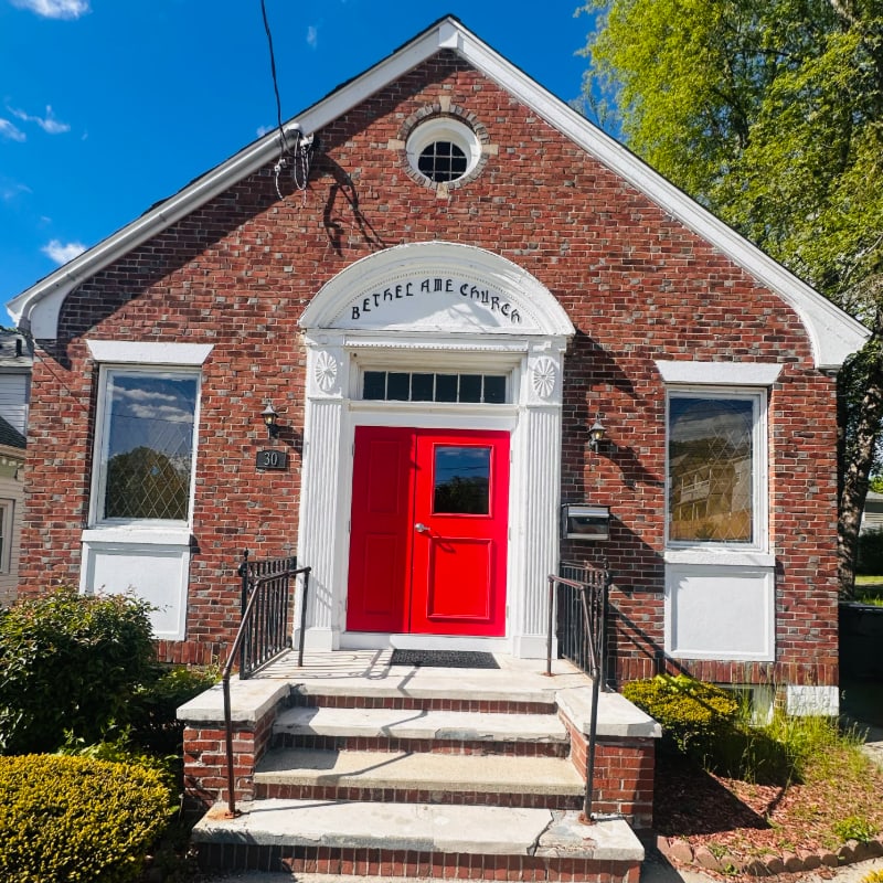 A bright red door stands out on a brick church with a clear blue sky and green trees in the background.