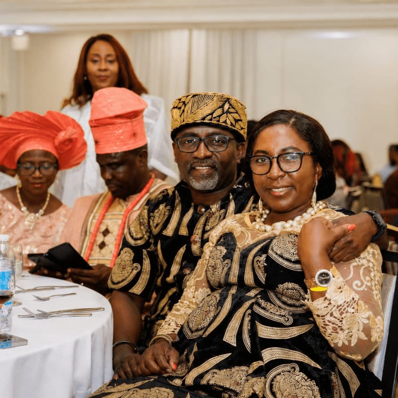 A smiling couple in traditional attire poses in the foreground, while other guests in festive clothing are visible in the softly lit background of a formal event.