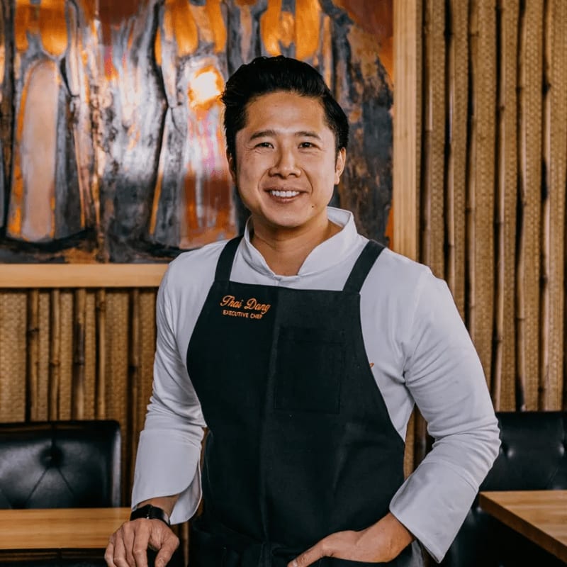 A smiling man in a chef's coat and apron stands in front of a textured wall with artwork and bamboo accents.