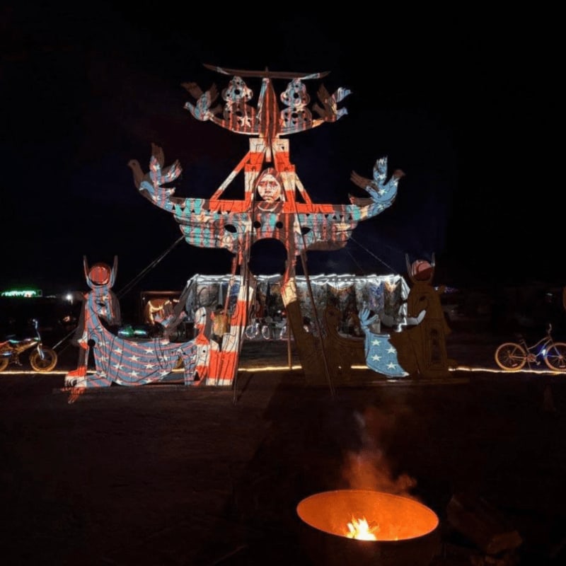 A large, illuminated art installation with Native American motifs stands in the dark desert, with a campfire burning in the foreground.