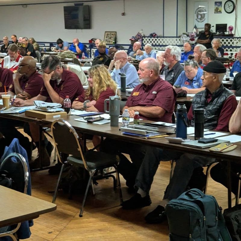 A group of people are seated at tables in a room, with some in the foreground looking towards the front and others in the background attending a presentation.