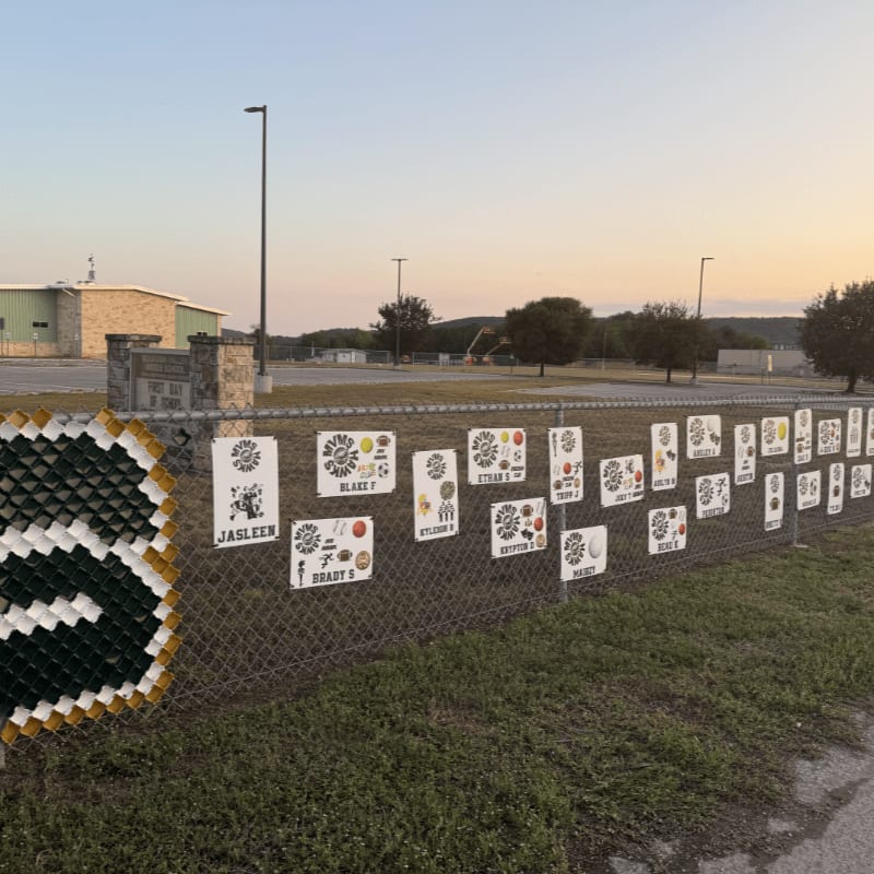 A chain-link fence adorned with numerous student artwork squares and a large green and yellow "S" decoration stands in the foreground, with a building and trees under a pale sky in the background.