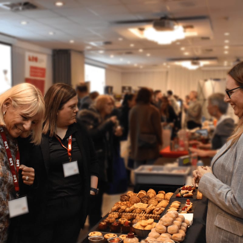 In the foreground, two women examine a table laden with pastries, while in the blurred background, a crowd mingles in a brightly lit room.
