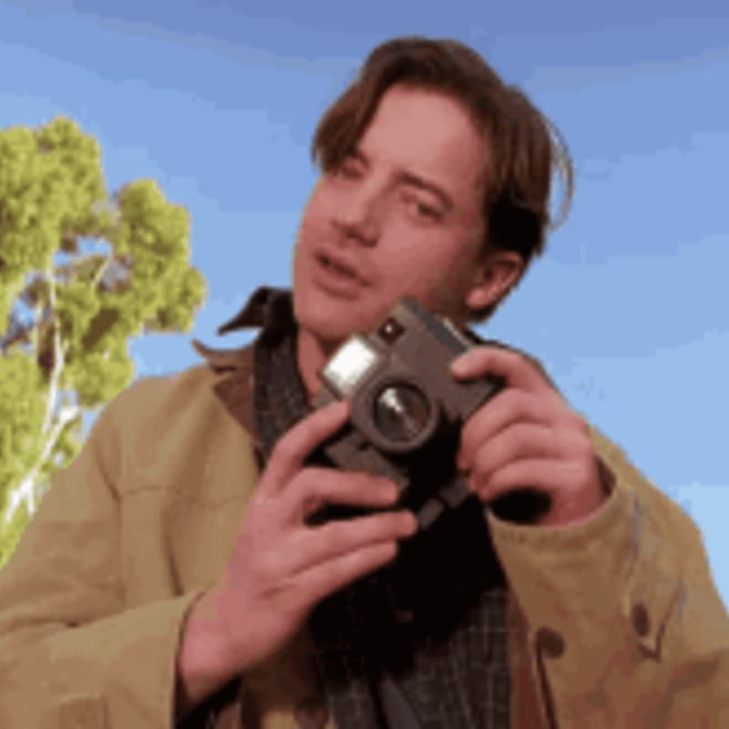 A young man in a tan jacket holds a camera in the foreground, with a bright blue sky and a green tree in the background.