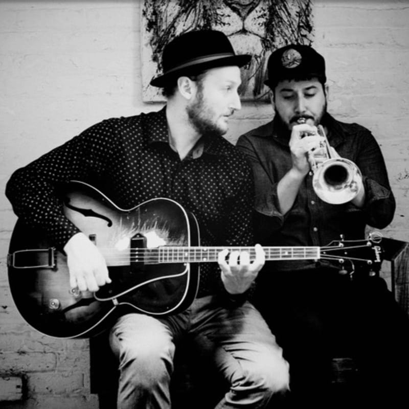 A man in a hat plays a guitar in the foreground while another man plays a trumpet in the background, with a lion artwork on the wall behind them.