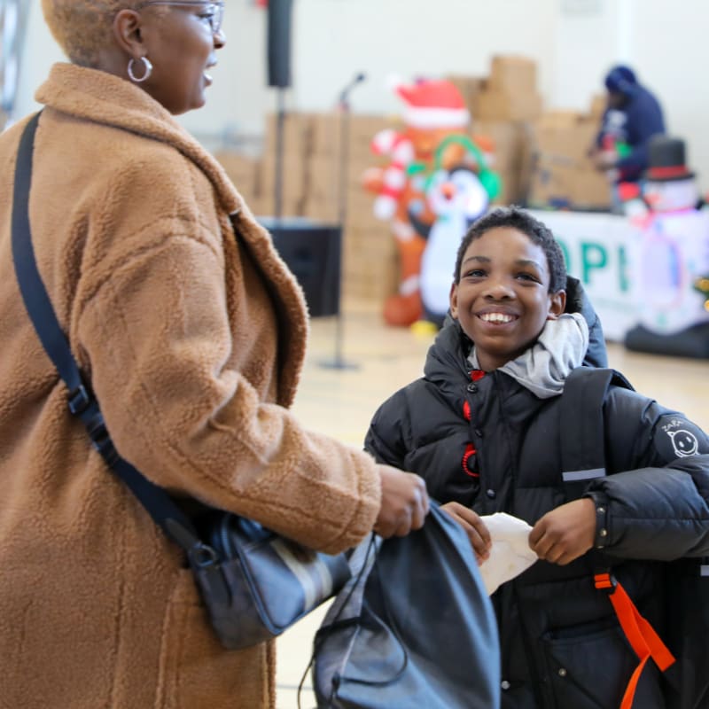 A smiling boy in a puffer jacket receives a bag from a woman in a brown coat, with festive decorations and people in the background.