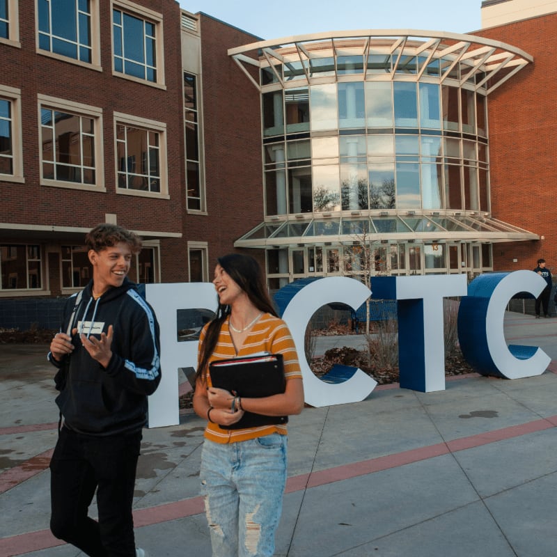 Two smiling students walk past large white letters spelling "FCTC" in front of a modern brick building.