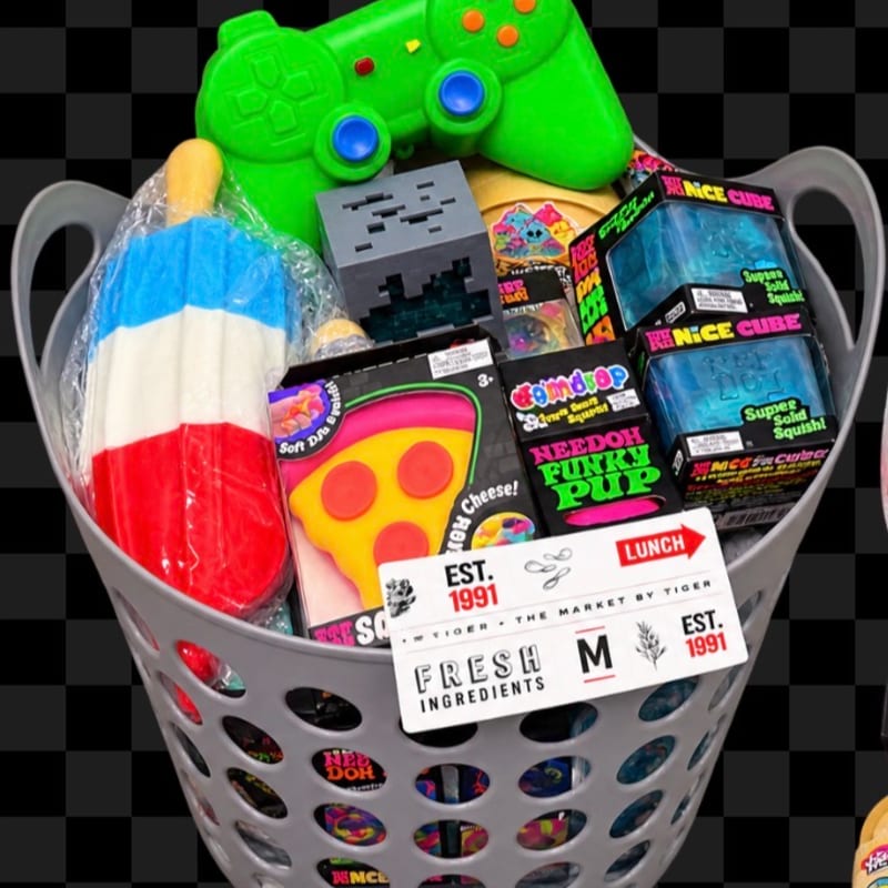 A gray laundry basket overflows with colorful toys and snacks, set against a black and white checkered background.