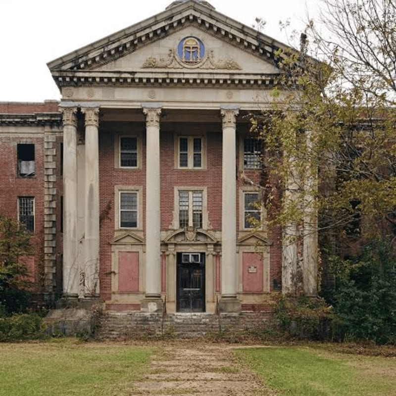 Overgrown grass and a path lead to the imposing, dilapidated brick facade of a grand building with large columns and boarded-up windows, set against a muted, overcast sky.