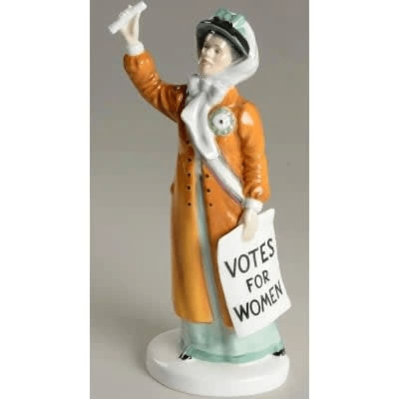 A porcelain figurine of a suffragette in an orange coat and green hat stands on a white base, holding a "Votes for Women" sign and a scroll against a plain gray background.