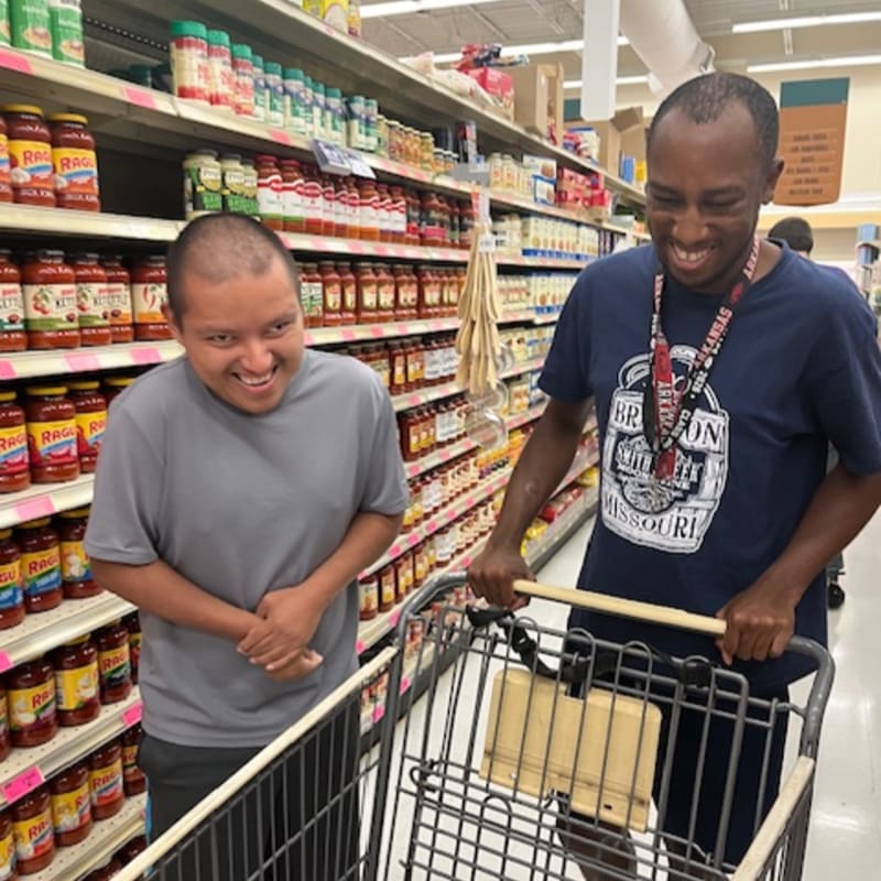 Two smiling men push a shopping cart down a grocery store aisle stocked with jars of sauce.