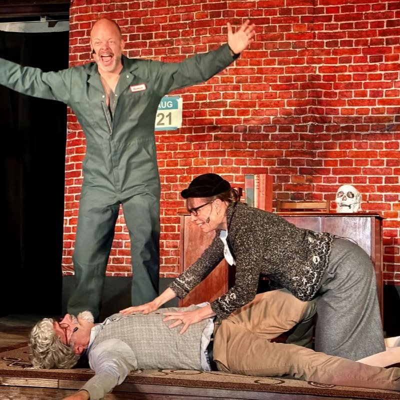 A man in a green jumpsuit stands with arms outstretched in front of a brick wall, while a woman kneels over a man lying on the floor.