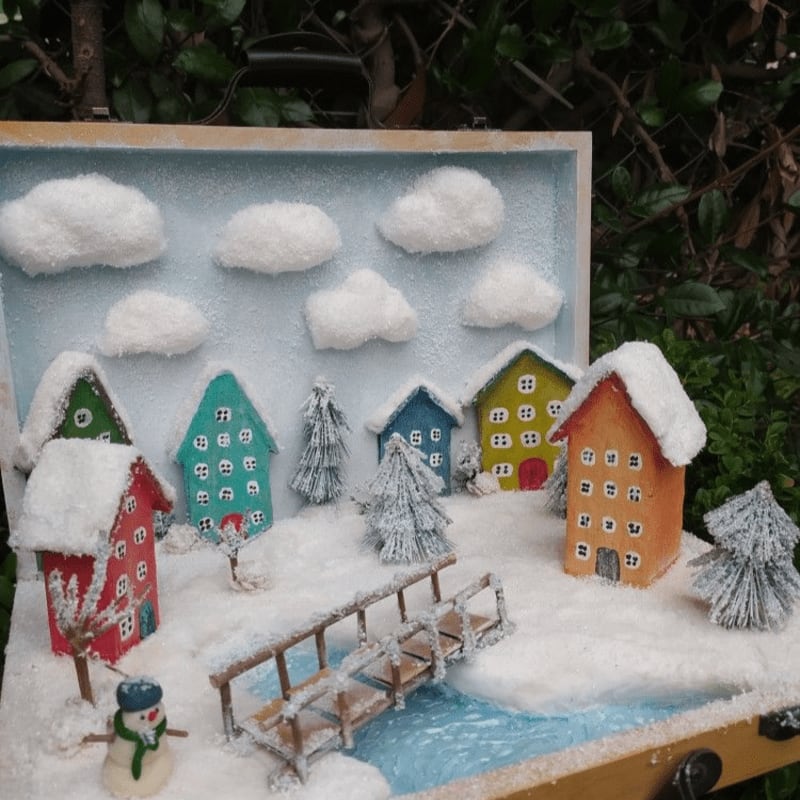 A miniature snowy village with colorful houses and a snowman in the foreground sits against a backdrop of fluffy clouds and greenery.